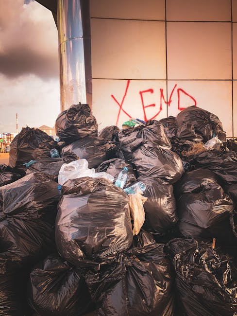 A large pile of black rubbish bags, some torn open revealing plastic bottles and waste inside, is stacked against a building with a tiled exterior. The bags are placed on a paved surface in an outdoor