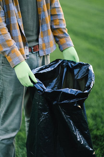 A waste management worker dressed in a yellow and red uniform is positioned behind a large red rubbish collection truck, handling debris at the back of the vehicle on a paved roadside. The truck, desi